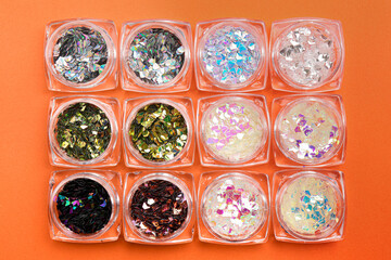 Jars with colorful shiny glitter on orange background, flat lay