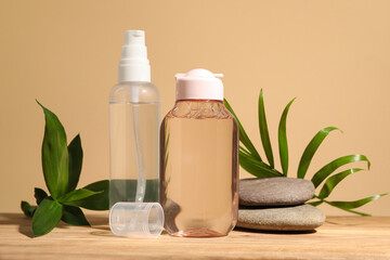 Fototapeta na wymiar Bottles of micellar water, green leaves and spa stones on wooden table against beige background
