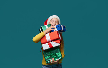 Сheerful child girl in Santa Claus hat holds a red gift boxes  on a turquoise background. Preparing for the Christmas holidays. Season of winter holiday sales and discounts