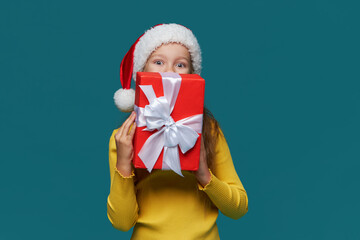 Сheerful child girl in Santa Claus hat holds a red gift box on a turquoise background. Preparing for the Christmas holidays. Season of winter holiday sales and discounts