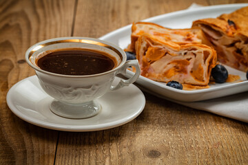 Apple strudel on white ceramic plate and cup of coffee on wooden table