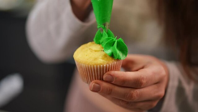 Close-up female hand squeezing green icing from piping bag on baked cupcake in slow motion. Young unrecognizable plus-size Caucasian woman decorating delicious pastry indoors