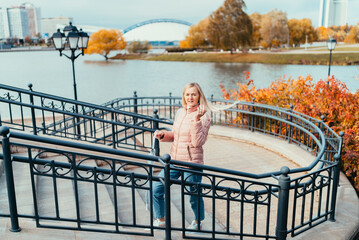 A blonde girl goes up the stairs on the lake against the background of autumn leaves and laughs on a sunny day