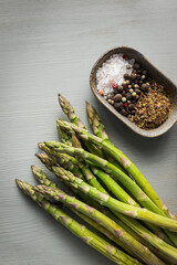 Closeup view of a bunch of green asparagus and a ceramic tray full of spices, salt and pepper balls over a wooden table.