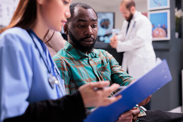 Close up of african american man reviewing medical records along with asian female member of in house hospital staff. Doctor indicating treatment to young male patient at sanatorium lobby. - obrazy, fototapety, plakaty