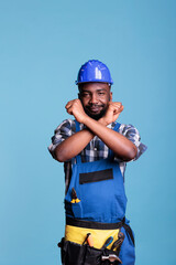 Disapproving african american builder making stop gesture with crossed arms while standing on blue background. Construction worker showing deny hands symbol while refusing to accept more work.