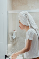 After shower body and head wrapped in towel woman looks in mirror touches moisturized soft healthy face skin feels satisfied enjoy spa cosmetics treatment procedure, morning care hygiene.