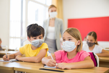 Portrait of small school girl and boy in protective masks sitting together in classroom during lesson in elementary school