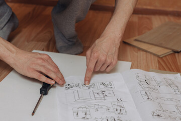 From above of crop man reading paper assembly instruction while assembling furniture on floor in room in apartment 