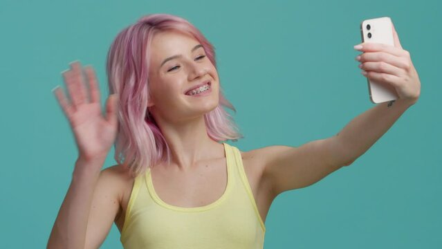 Young woman with pink hair on pastel background. Millennial travel blogger waving and sending air kiss to camera on smartphone. Make video for social media or memories. Generation z new concept studio
