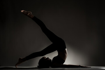 Fit ballerina girl lying and stretching practicing yoga poses. Side lit silhouette against dark background.. .