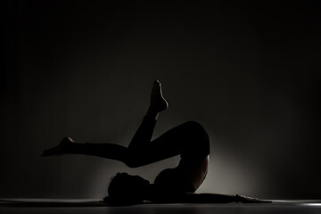 Fit ballerina girl lying and stretching practicing yoga poses. Side lit silhouette against dark background.. . © Nikola Spasenoski