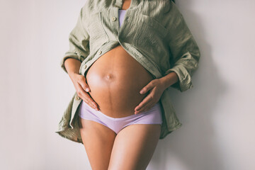 Pregnancy belly closeup. Pregnant woman wearing underwear and casual cotton shirt at home relaxing holding expecting tummy for skincare, health, lifestyle - obrazy, fototapety, plakaty