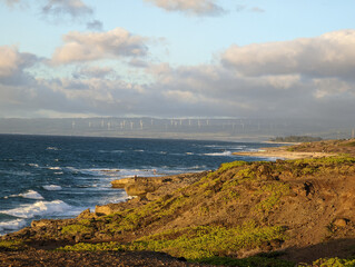 This is the view from the western point of O'ahu looking back east. I was in Ka'Ena Point Nature reserve and was looking bake east over the coast and saw the windmills lighted in the setting sun.  - obrazy, fototapety, plakaty