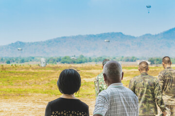 Back view of Asian parents look with worry and concern during parachute training from airplane for army cadet with blurred image of parachute and landscape in background. Family relationship concept.
