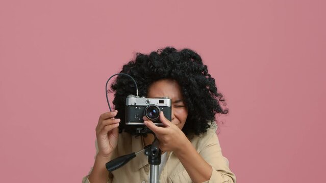 Waist up portrait of focused African American woman looking at camera and viewfinder. Photographer posing with analog camera on tripod standing in bright pink photo studio on isolated background
