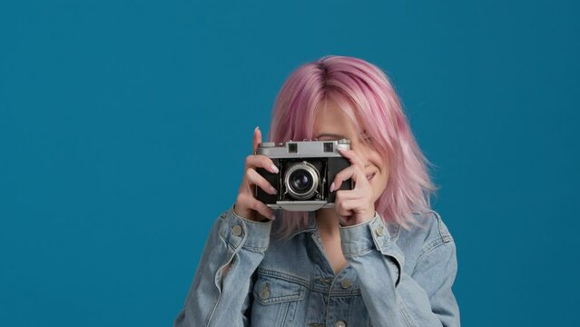 Hipster girl with nostalgic analog film camera. Young professional female photographer with pastel pink hair color taking photos in photo studio bright blue background. Creative industry jobs concept