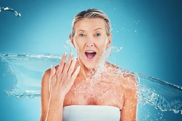 Poster Beauty splash, senior woman and wow face in studio for cleaning, fresh and grooming on blue background. Skincare, water splash and elderly lady model shocked by water, hydration and moisture product © Allistair F/peopleimages.com