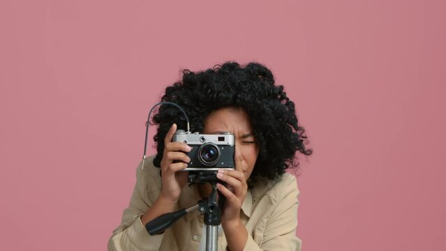 Waist up portrait of focused African American female photographer posing with analog camera on tripod standing in bright pink photo studio on isolated background, looking at camera and viewfinder