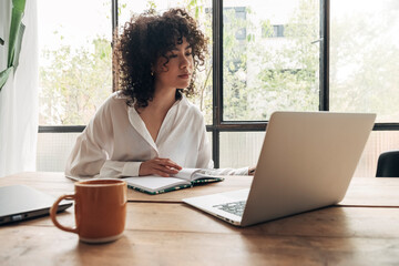 Young pretty mixed race woman studying at home with laptop. Taking notes in notebook. Bright spacious living room.