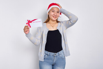 Thinking and pensive young Asian woman in a Santa Claus hat holding a gift certificate coupon...