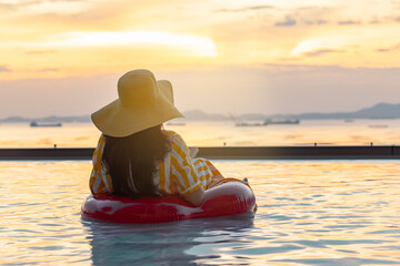 
Back side of Happy traveler asian woman floating with red rubber inflatable ring in infinity pool, looking out to sea, watching sunset serene near sea beach in vacation.