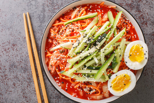 Korean cold noodles with spicy sauce, topped with cucumber, boiled egg and crushed sesame seeds in a black bowl closeup. Horizontal top view from above