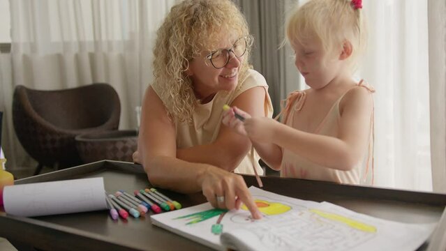Grandmother teaches her granddaughter to draw with pencils in a coloring book. The concept of communication in the family and learning at home with pleasure