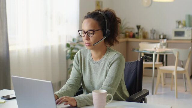 Medium shot of woman with headset sitting in wheelchair and typing on laptop while working as call center agent from home