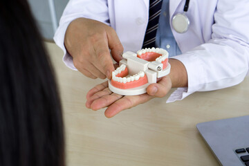 Dentist in white gown and stethoscope pointing finger at tooth model, explain to young patient about orthodontics and braces. Help reduce gum pain. Healthcare and medicine concept. Closeup