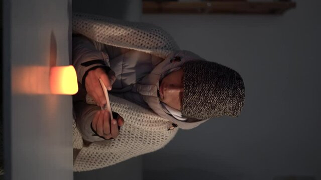 Arabic man dressed in a warm winter hat sits at home at a table and warms his hands from a burning candle. Concept of no heating in winter at home.