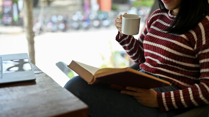 Bright Asian female reading a book while relaxing sipping coffee in the cafe. cropped image