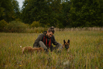 Naklejka na ściany i meble Male pet owner smiling, sitting in green grass and stroking Australian and German Shepherd with hands. Young Caucasian man with dreadlocks and beard sitting in autumn field with two purebred dogs.