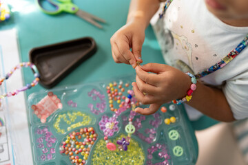 Little girl beads colorful braided necklace at table indoor.