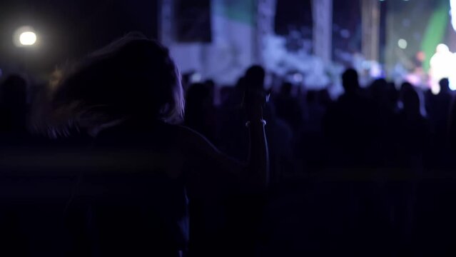 Silhouette of carefree happy woman is dancing at music performance held outdoors at summer night. Happy energetic lady is moving at open air concert in the darkness with stage lights on the background