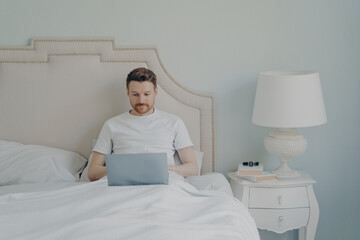 Attractive young european man watches video online on portable laptop computer while lying in bed
