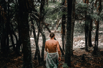 caucasian boy with bare back legs covered by rolled up towel walking towards river water between...