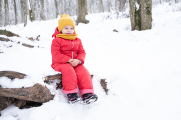 Fototapeta na wymiar Cute little girl in pink snowsuit plays with snow in winter forest