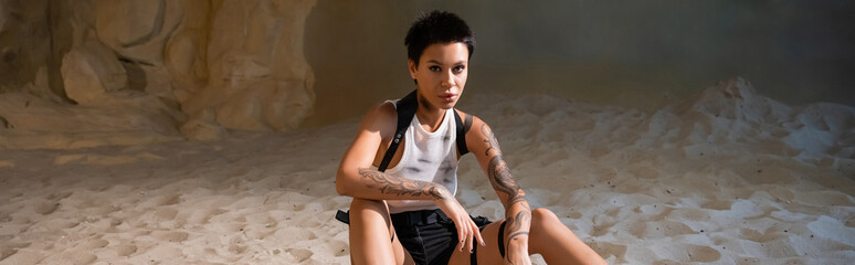 tattooed and sexy archaeologist in dirty crop top and shorts sitting on sand, banner.