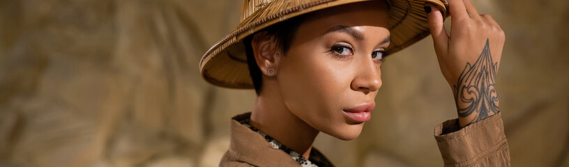tattooed archaeologist touching safari hat and looking at camera in desert, banner.