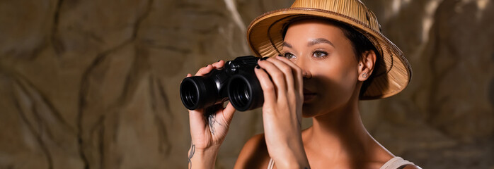young and pretty archaeologist in safari hat holding binoculars and looking away in cave, banner.
