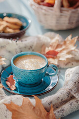 Autumnal composition with white knitted scarf, blue cup of coffee and dry yellow leaves on a table. Autumn mood.
