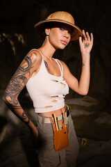 sexy tattooed archaeologist in tank top touching safari hat and looking at camera in dark cave.