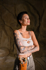 sexy and tattooed archaeologist standing with crossed arms and looking away in dark cave.