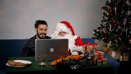 Santa Claus is sitting next to the Christmas tree with a laptop. The guy with Santa Claus are sitting on the couch in front of a laptop.