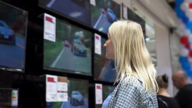 Young girl with white long hair buys new TV at electronics store. She chooses best model for her happy family home. many TVs or monitors stand on shelf or windscreen.
