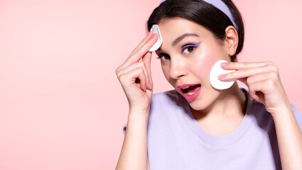 Modern excited amazed  girl removing makeup from face using cotton pads, apply cleansing cosmetics products to make skin clear on pink studio background. Skincare daily routine concept. © deniskomarov