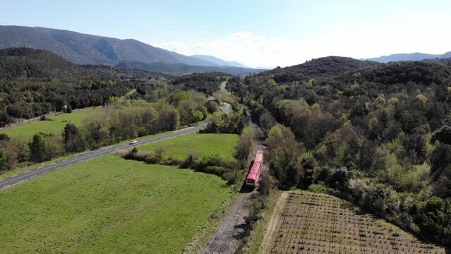 Aerial: red and white old train among vineyards and green fields by a road with light traffic in southern France