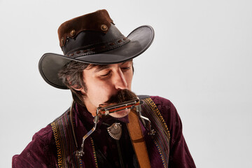 Portrait of man with moustaches in country style clothes, vintage hat playing harmonica isolated...