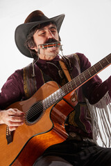 Portrait of man with moustaches in country style clothes playing guitar and harmonica, performing isolated over white background. Cowboy style